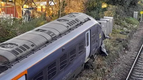 John Horton A blue train which has derailed. It is in undergrowth in front of three grey metal boxes