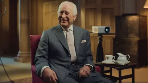 King's Foundation King Charles in a grey suit sitting in a chair, in a room where videos are being screened