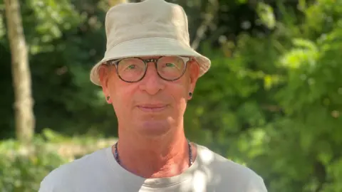 A man with glasses is wearing a bucket hat and a white shirt. He is also wearing a beaded necklace and black earring studs. He is standing in front of some trees. 
