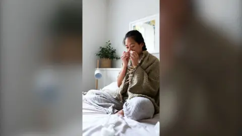 A woman suffering from a cold, blowing nose with paper tissue sitting on bed.