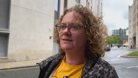 BBC/DAN BATER Emma Powell, who has blonde hair and is wearing glasses, with roads and buildings behind her.