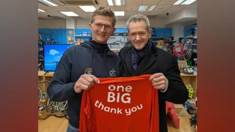 Michael Grisenthwaite Alexander Armstrong is on the right and has grey hair with a black coat and blue and white scarf. He is holding one side of a red top that reads "one big thank you" in white writing. On the left is Michael who has brown hair and glasses and is wearing a blue navy hoodie, and is holding the other side of the top.