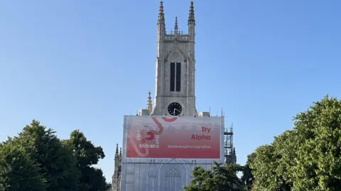 The church tower which has a clock just above the scaffolding, which is around the tower