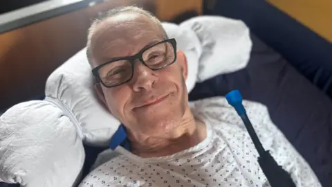 A man with short grey hair lying in a hospital bed with black glasses on and smiling up at the camera. He has his head on a pillow and is wearing a white hospital gown with blue dots on it.