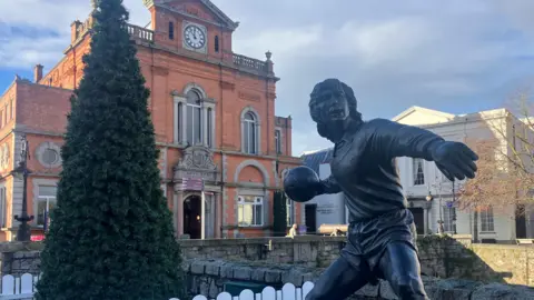 The image shows a statue of footballer Pat Jennings, which stands outside the theatre in Newry. A christmas tree is behind the statue.