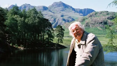 Alfred Wainwright at Blea Tarn. He has white hair and glasses and is wearing a beige jacket. He is sitting in front of a small lake, with trees and fells in the background.