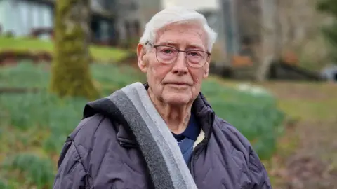 Alan Kelly outside Hawes End. He has short white hair and blue eyes, with glasses with a thin metal frame. He is wearing a grey coat and has a striped grey scarf over one shoulder. He is looking at the camera with a serious expression. Behind him is Hawes End, a former orphanage, in the blurred background.