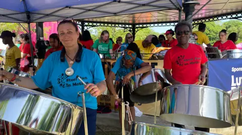 Toby Friedner/BBC A steel band performs under a tent within a park during a carnival celebration. They wear matching blue and red T-shirts that have the words "steel pan in motion" on the front.