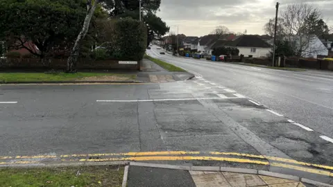 Herbert Avenue, a single carriageway main road with houses on both sides, has a turning lane into Manor Road, seen on the left.