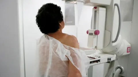 Reuters A woman with short, dark, curly hair wears a fabric hospital gown and stands in front of a breast screening (or mammogram) machine, which is a large, white-coloured piece of medical equipment