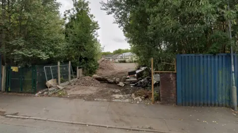 Trees and bushes are to the left and right of an entrance. Part of a road and pavement are at the bottom of the photo. 