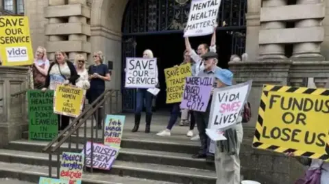 A group of adults holding banners and placards stand on the steps of an official looking building
