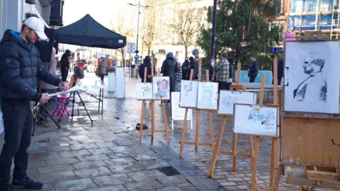 Asylum Artist Quarter CIC A man standing on a rainy street holding at art work. He is wearing a puffer jacket and a cap. In front of him are drawings on easels. The backs of people walking by can be seen as well as what looks like a Christmas tree which has not yet been decorated. 