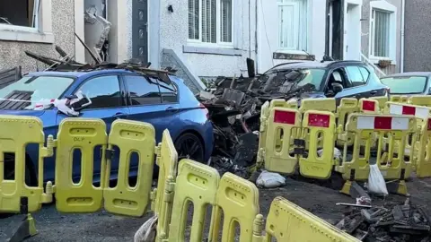 A fenced-off area of street with yellow barriers around damaged pavements. There are two cars which have bits of wood and rubble on their roofs and bonnets. Some of the wood is blackened. 