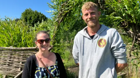 Shariqua Ahmed /BBC Julie wearing a black cardigan and a blue and yellow patterned top standing on the left and Lewis wearing grey zipped hoodie standing on the right- in front of green trees