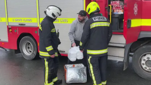 A man hands over to plastic carrier bags with food inside them to two firefighters. They are all stood next to a red fire engine.