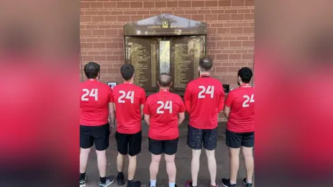 Erica Dillon Five male runners wearing red running tops with the number 24 on the back standing in front of Liverpool FC's eternal flame memorial to those who died in the Hillsborough disaster.