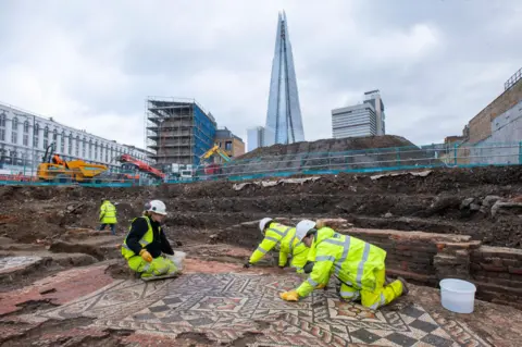 Mola/Andy Chopping Four archaeologists wearing hard hats and hi-viz clothing are bent down or on all-fours as they work on a Roman mosaic. The Shard is in the background of the photo