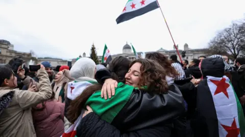 Reuters Two women - one wearing a Syria flag around her shoulders - embrace at a celebration to mark the end of Assad's regime at London's Trafalgar Square on an overcast December day 