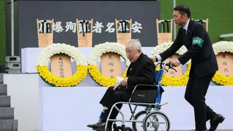 STR/JIJI PRESS/AFP via Getty Images Bomb survivor, Hiroshi Nishioka, who is very elderly and in a wheelchair, is pushed by another man, while wreaths of flowers are in the background.