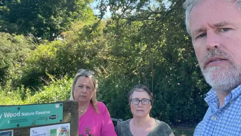 Supplied Liberal Democrat councillors Amanda Hopgood, Elizabeth Pears and Mark Wilkes stand next to a wooden board showing maps of Aykley Wood Nature Reserve. Amanda Hopgood has straight blonde hair and wears a bright pink top. Elizabeth Pears has short dark hair, glasses and a grey top. Mark Wilkes has white beard and hair. He wears a blue and white checkered shirt. 