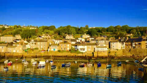 A picture of Mousehole. There are houses on the harbour. In front of the houses are small boats moored in the water. The sky is blue. 