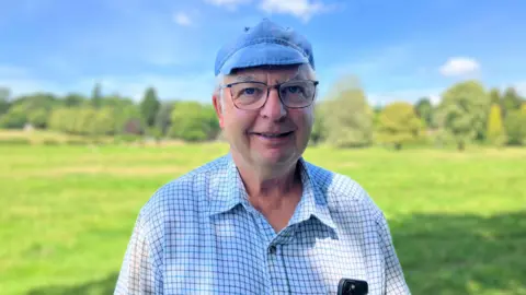 A man stood in a field. he is looking at the camera smiling wearing a blue milking pallor cap, glasses and a check shirt.