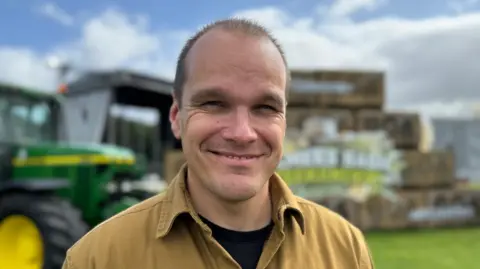 A man is wearing a brown shirt over a black t-shirt. He has short black hair. The background is blurred but a green tractor can be seen.