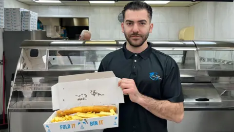 Chris Onisiforou is pictured standing in a fish and chip shop. He is holding a portion of fish and chips in an box open. He is wearing a black polo shirt and he has short black hair which is gelled back and a short beard. 