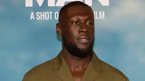 Stormzy at the Big Man premiere. He has a short beard and shaved head and wears a mossy green shirt. He's pictured in front of a board printed to resemble a blue sky. 