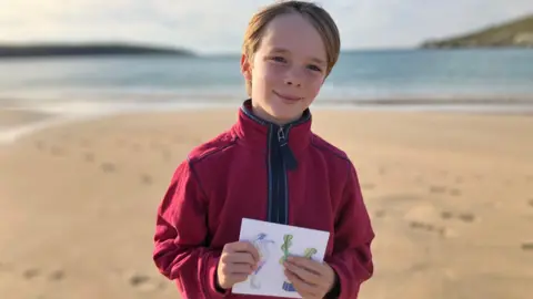 A young boy with blond hair and a red fleece stands of a beach in the sunshine, hoping an illustrated greetings card 