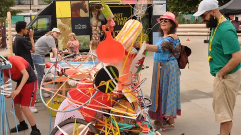 A quirky art piece made up of colourful objects such as tennis rackets and hoola hoops, pieced together. Two people are admiring the piece, a woman in a long blue dress with sunglasses and a pink hat and a man in a green top and beige shorts with a grey baseball cap. The art piece is outside, other activities are taking place in the background. 