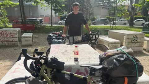@devonbaistobyronbay A table with graffiti on. There is a bike in front of the table. Behind the table is Conrad holding his bike up. He is wearing a brown top and grey shorts and a cap. Behind him are trees and parked cars. 
