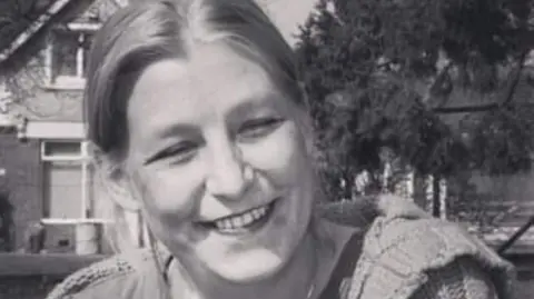 Dawn Sturgess - a black and white image, where Dawn is sitting outside with a house and trees behind her. She has light hair and is smiling into camera.