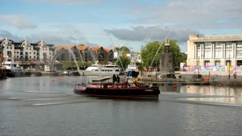 A black boat with red stripes sailing in Bristol Harbour. There are buildings in the distance.