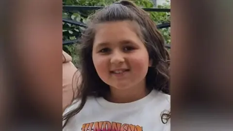 A young girl, with long brown hair, wearing a white t-shirt. She is smiling at the camera. 