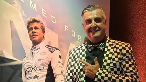 Martin Donnelly Martin Donnelly is standing on the right looking towards the camera smiling He is wearing a black and white chequered suit jacket, and a black shirt with a tie to match. He is point to a poster of the film which has a picture of Brad Pitt wearing a white racing suit.