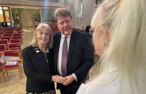 Sue and Alex Tatham in the church facing the camera, with empty red seats behind them. There is a light green wall at the back and a white wall to the right.