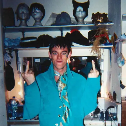 Luke Evans Luke Evans in a dressing room, wearing some make-up. He has a blue jacket on and black trousers. He has got his middle finger on both hands up at the camera