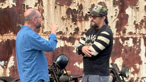 Jason Momoa standing talking to an Electrogenic staff member. Next to them is a vintage motorcycle.