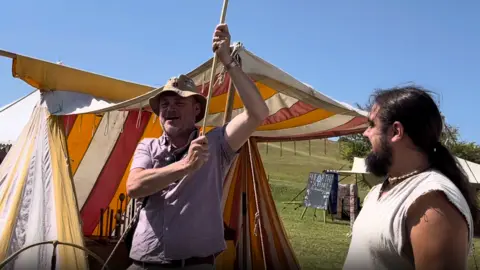Al Murray stands in front of a striped, colourful marquee in a field. He is holding up a carnyx - a historical horn - with only its pole visible. Another man with long hair in a ponytail watches on.