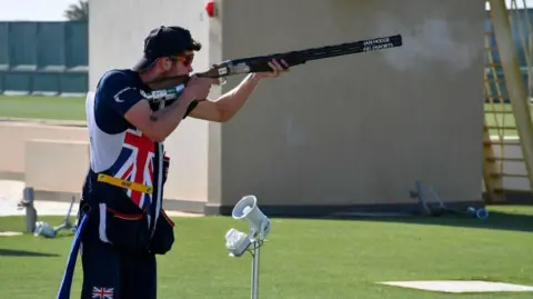 ISSF Grose is shooting at a target. He is wearing red sunglasses, the GB team uniform which features a Union Jack flag design and a cap which is on backwards. He is holding a long barrelled gun and there is smoke coming from the barrel.
