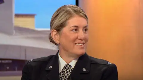 A close-up shot of Katy Sigley, wearing police dress uniform, sat on the BBC Breakfast red sofa. Behind is a studio backdrop which features images of Salford Quays.