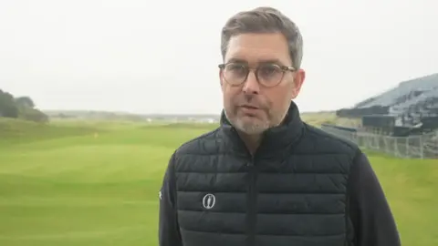 Mark Darbon, who is wearing a black gilet and jumper, is standing in the middle of a golf course. A viewing platform can be seen on the right-hand side. 