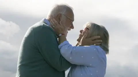 Curious North Productions An elderly couple, both with grey hair. Holding each other's faces. The man is wearing a grey jumper and has a beard, the woman is wearing a blue polka dot top. They are gazing into each others eyes, with a sky pictured behind them.