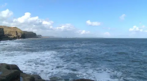 The sea laps rocks on a sunny day in Yorkshire; a grass-topped headland in the distance.
