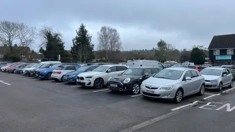 Holly Nichols/BBC An outdoor car park with rows of parked vehicles. The car park is bordered by leafless trees and evergreen trees, and beyond them are low houses and a gently sloping landscape. To the far right, part of a dark‑roofed building with shopfronts is visible. 