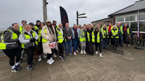 Emily Laidler A group of about 30 people, men and women, and a mix of ages but mainly in their early 20s about to set off on a charity walk. Some are wearing hi-viz jackets and one person at the front is carrying a collection bucket. They are standing on a pavement, with a cafe building to the right, and the sea can be seen behind them.