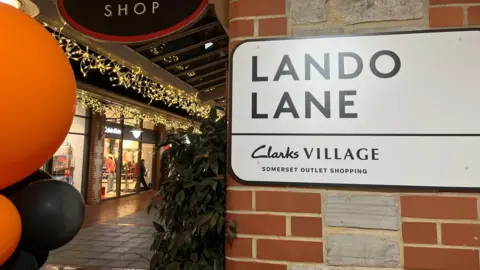 A sign saying Lando Lane on a wall in the clarks retail village in Somerset, it is a covered pedestrianised walkway and there are yellow and black balloons to the left of the shot