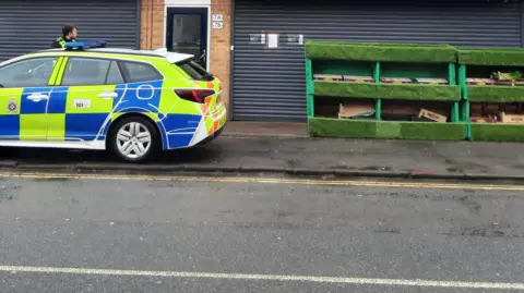 Wiltshire Police A police car parked by a shop which has its shutters down. There are empty green vegetable shelves out the front of the shop. A police officer is standing next to the car on the pavement.